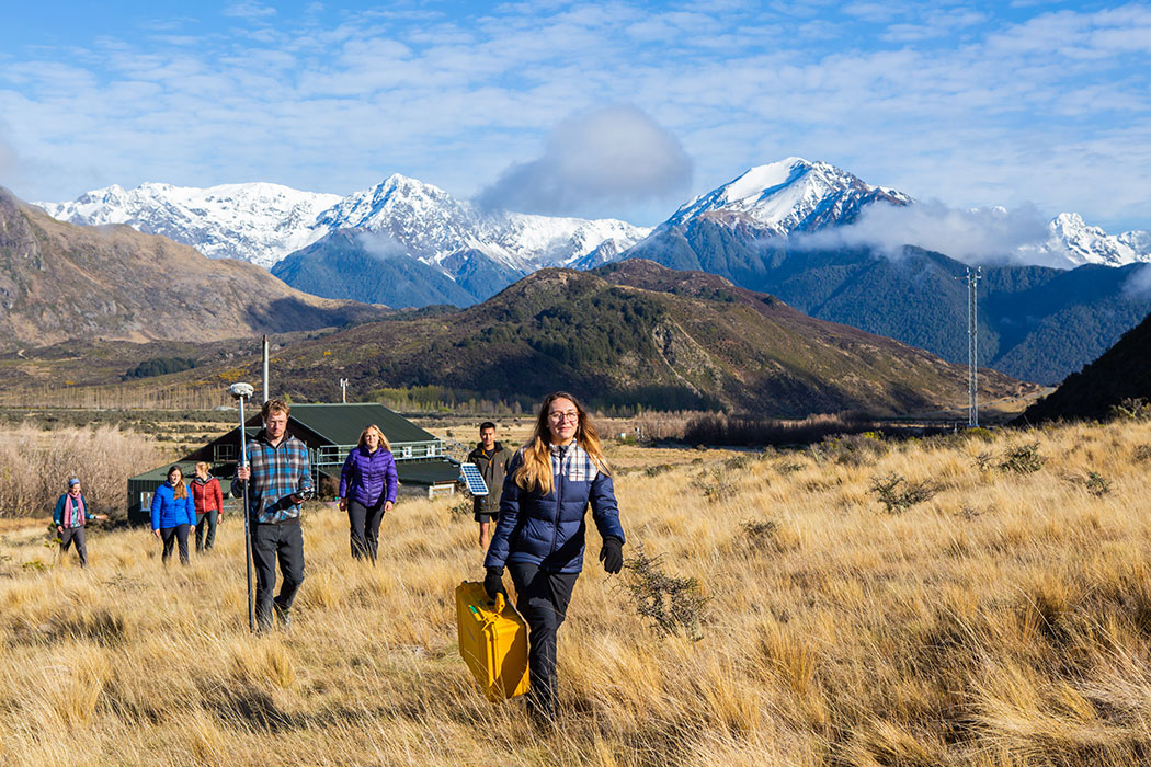 UC student researchers walking across the grassy plans of Cass carrying various equipment. Cass Field Station is behind them, and in the distance are snowy mountains.