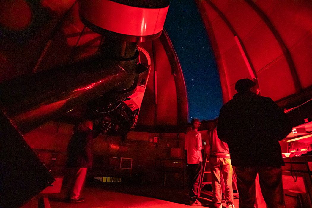 Interior of Mt John Observatory at nighttime, lit with an infrared light as people look through the telescope and through the roof opening to the night sky.