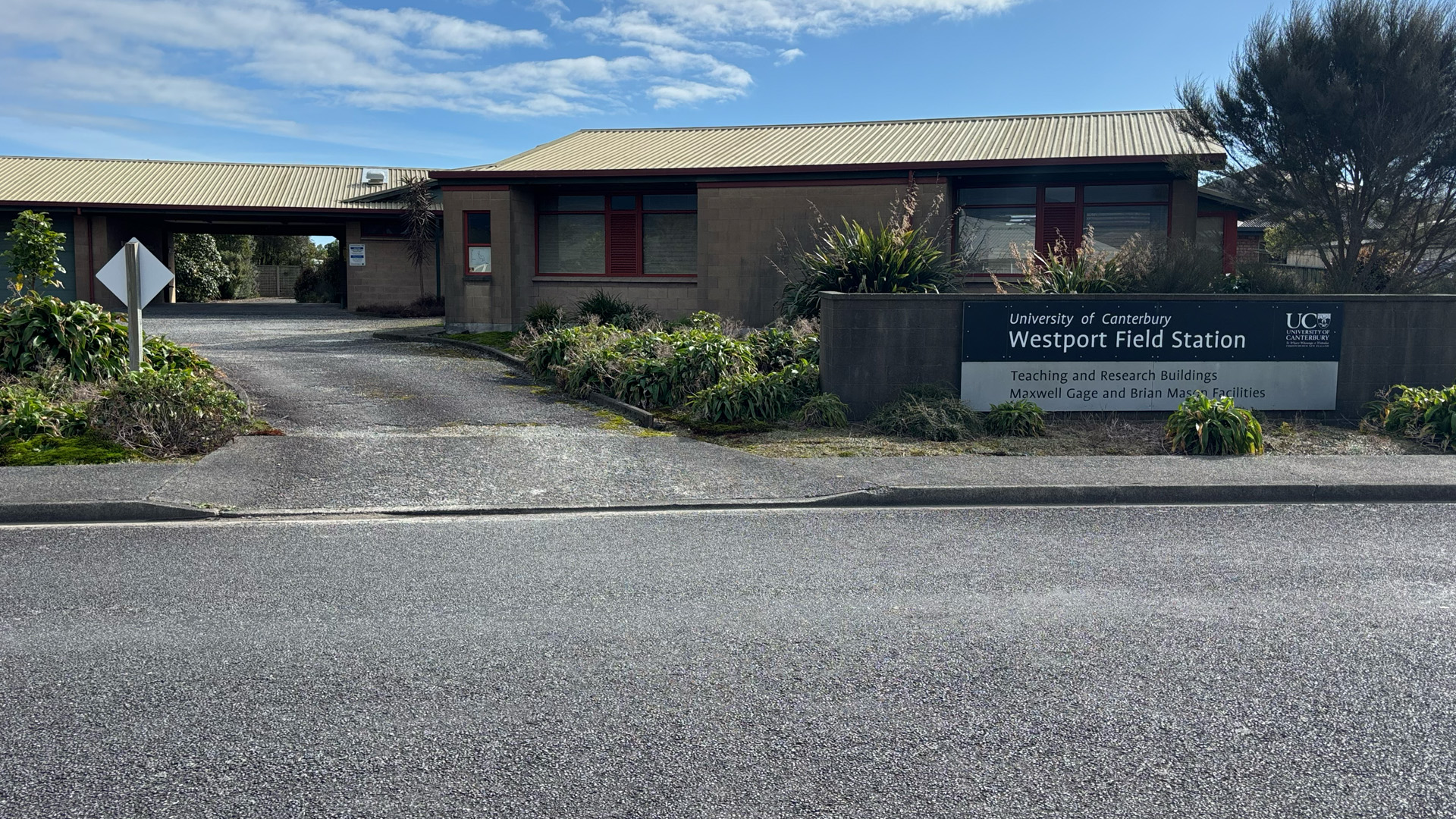 Small brick building facade on a sunny day with a driveway and sign reading "University of Canterbury Westport Field Station, Teaching and Research Buildings".
