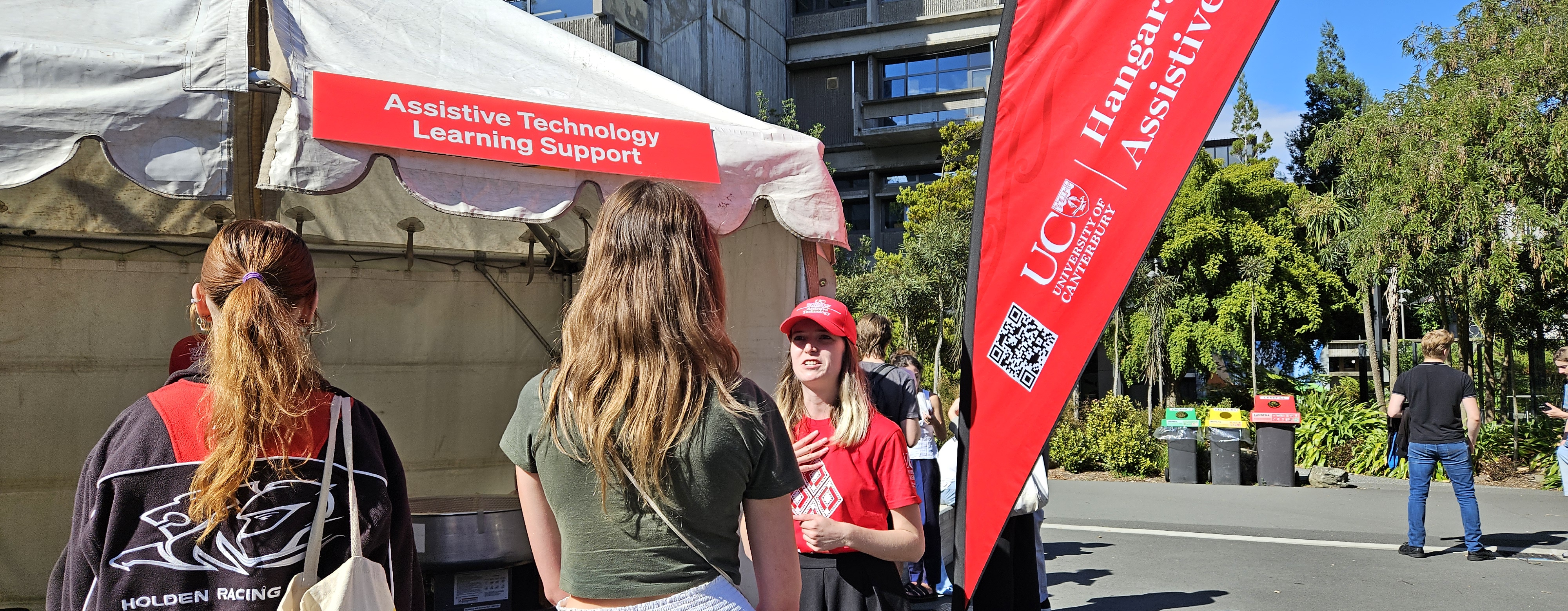Two Open Day visitors talk to a UC staff member standing beside a white event tent with the sign "Assistive Technologies Learning Support".