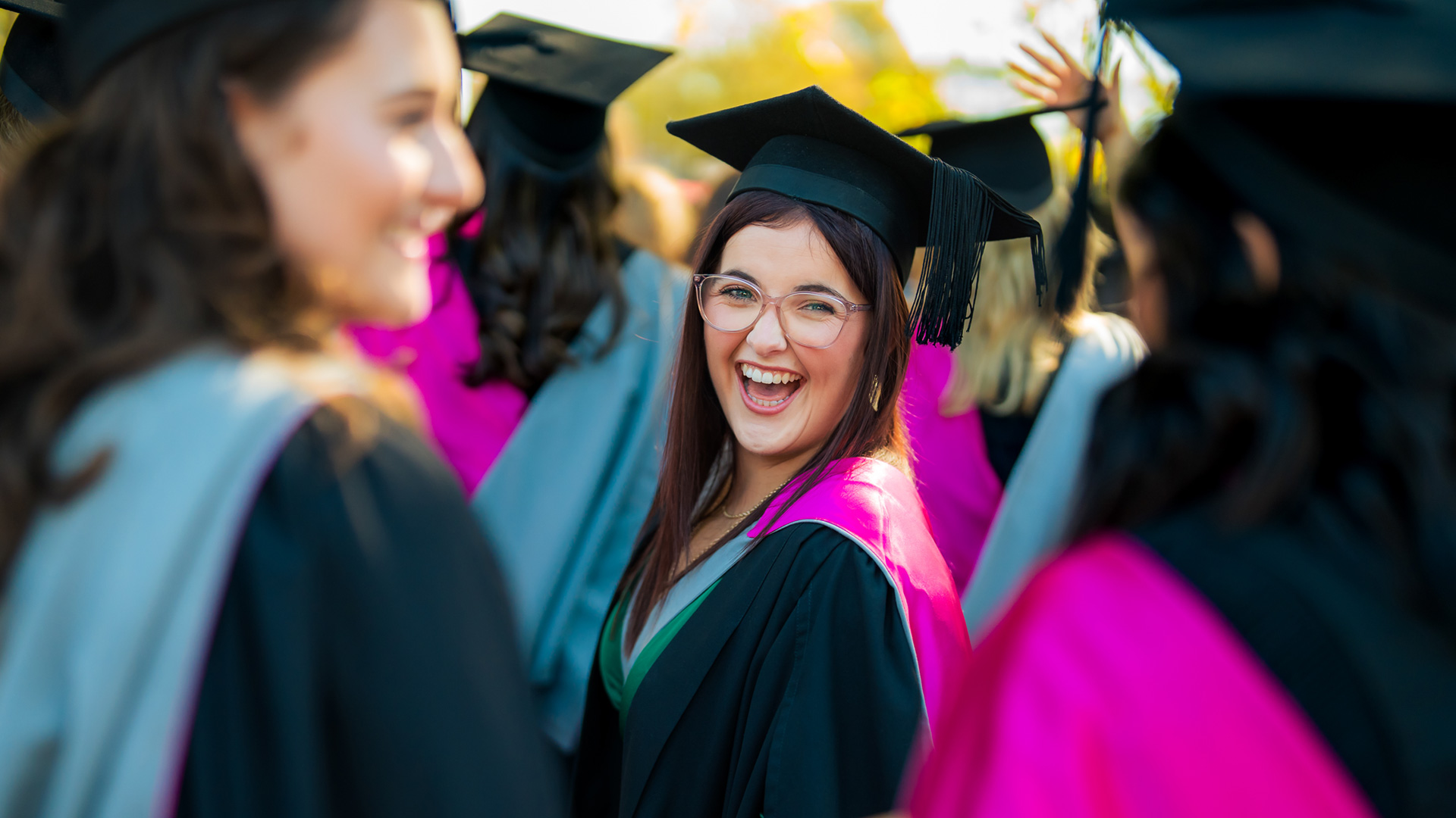 A UC student standing and smiling amongst a crowd of graduates, all wearing full graduation cap and gown regalia with bright pink hoods indicating their degrees in Speech and Language Pathology. 