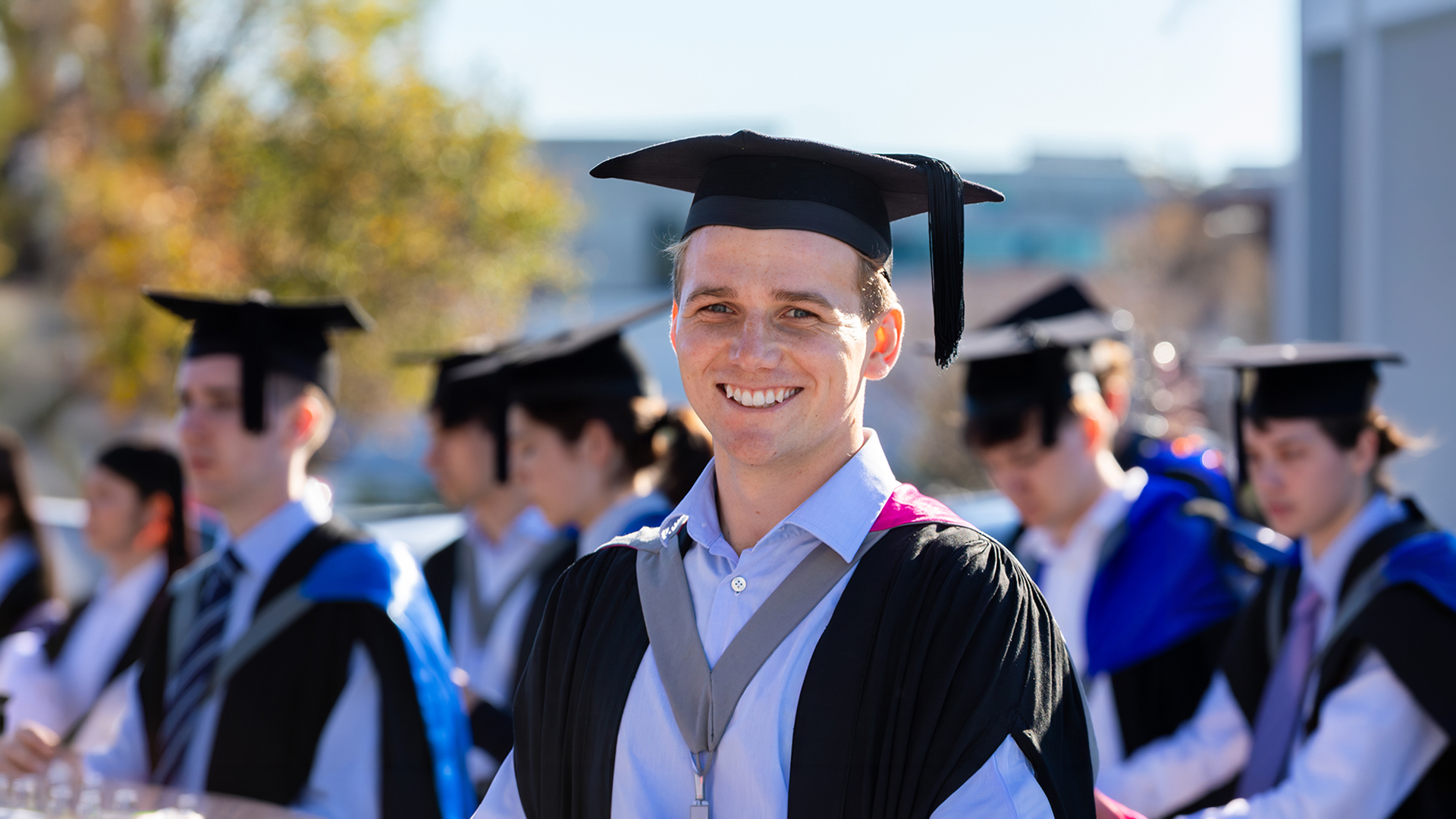 Smiling student wearing a graduation cap and gown, with other graduations seen walking in the background.