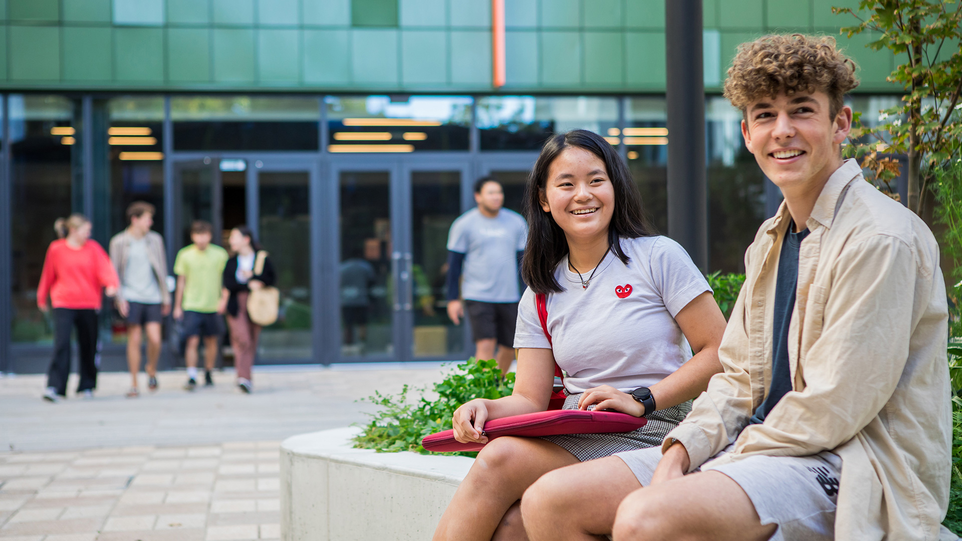 Two students sit by a garden bed outside Tupuānuku accommodation hall while other students walk through the courtyard behind them.