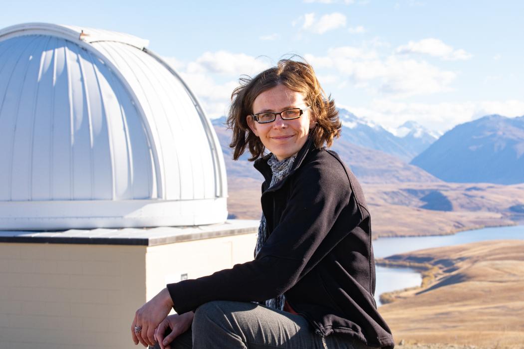 Dr Michele Bannister sitting outside the Mt John Observatory on a sunny day, with snowy mountains in the distance..