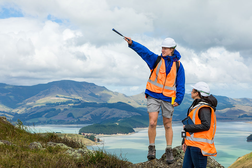 UC student Alex pointing and talking with another student at Rāpaki Rock on the Port Hills, overlooking Lyttelton Harbour.