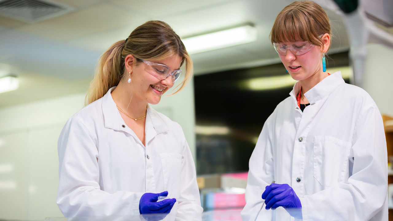 Two Chemical Formulation Design students wearing labcoats and gloves working in a lab at UC.