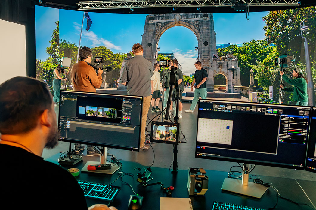 UC students using Virtual Production technology to film a scene on the LED volume. One student sits at a computer setting up the virtual screen with a shot of the Bridge of Remembrance in the Christchurch CBD, while in the background other students set up lighting, cameras, and marks for the actors.