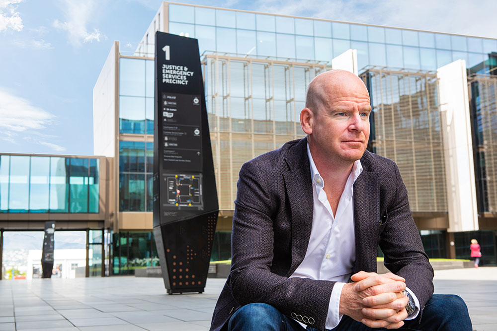 Person sitting on steps in a courtyard surrounded by modern buildings. A directions sign behind them reads "Justice and Emergency Services Precinct".