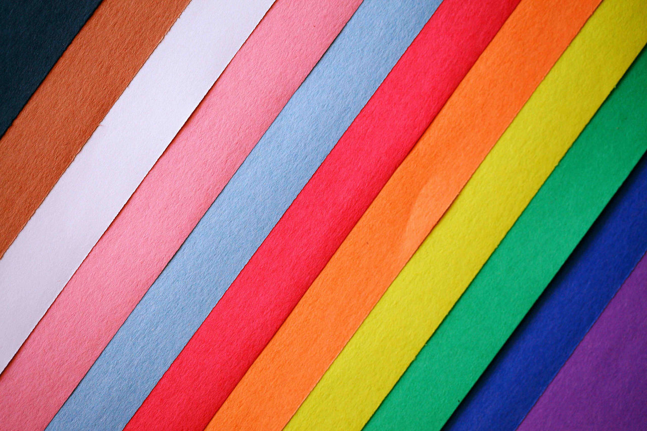 Strips of paper arranged diagonally down from the top left corner to the bottom right corner. In colour order; black, brown, white, pink, light blue, red, orange, yellow, green, dark blue, and purple to represent the progress pride flag.