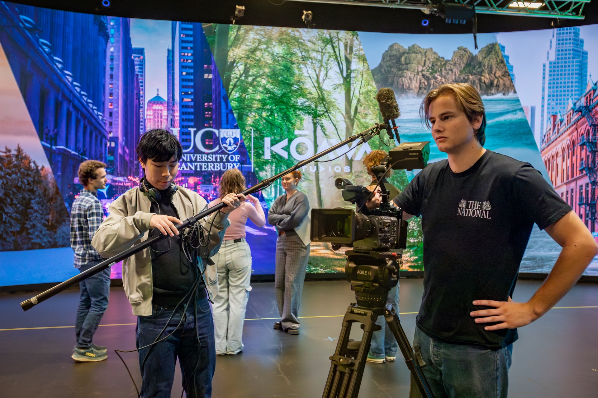 Students with cameras and other recording equipment standing in front of a large digital screen.