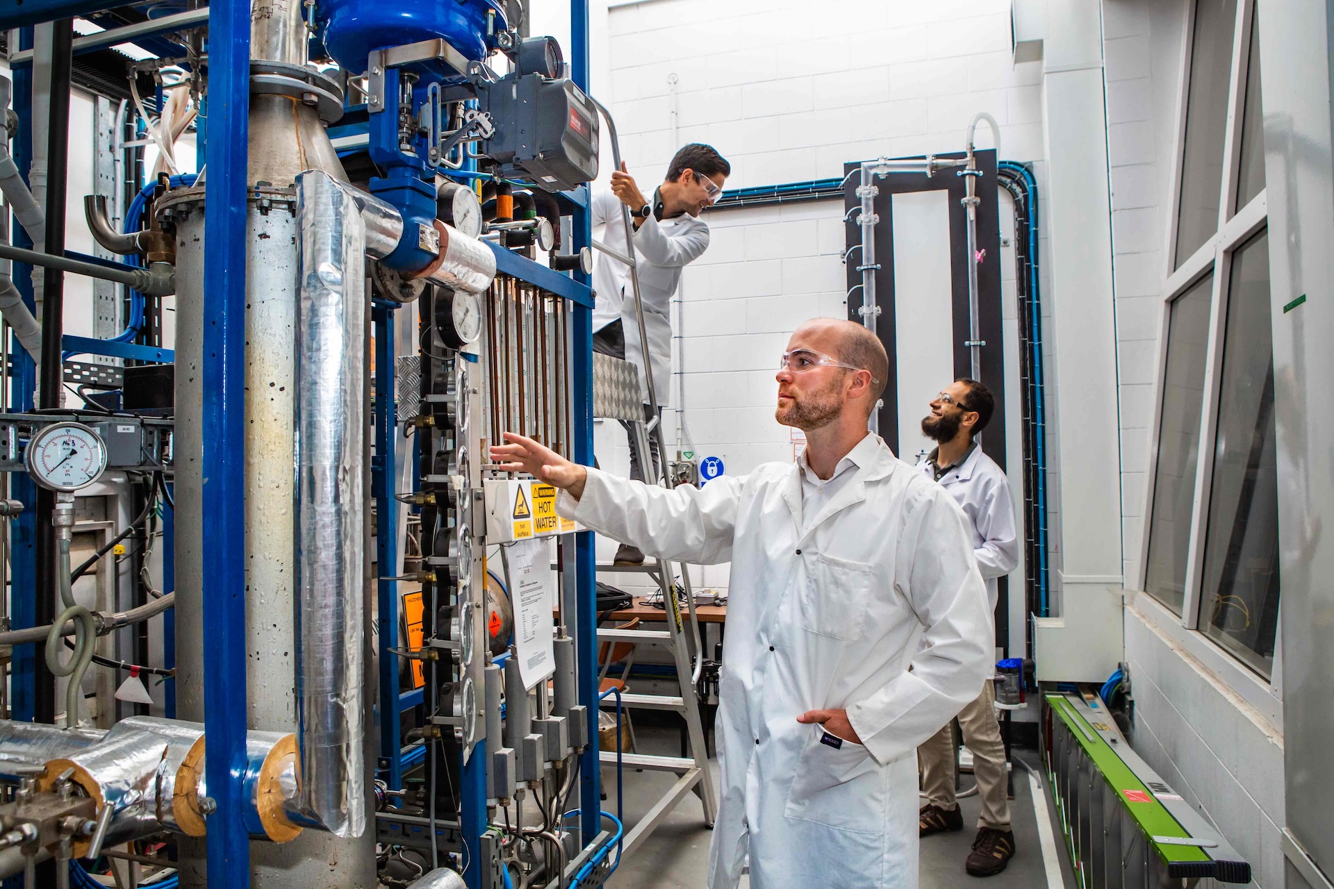 Three researchers wearing lab coats and safety glasses observing industrial pipe and gauge equipment