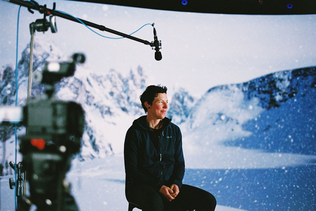A woman sitting in a sound stage on a stool with a virtual background behind her showing mountains with snow.