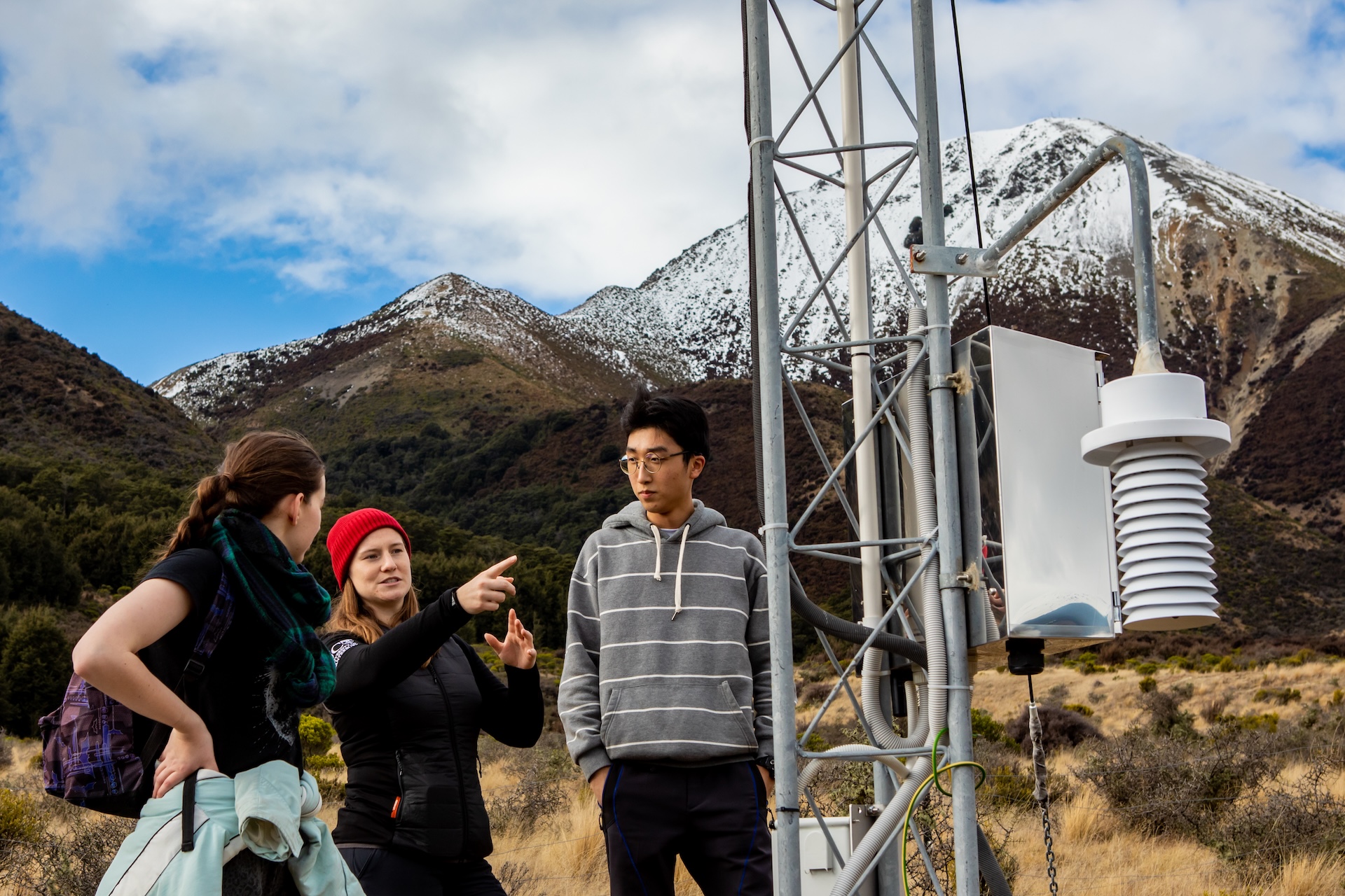 Three students standing next to and discussing aerial equipment with snowy mountains in the background.