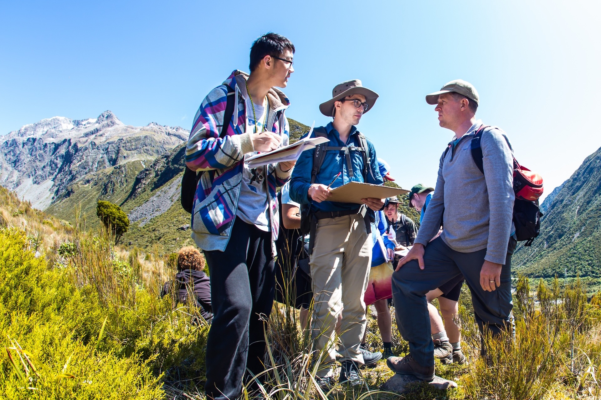 Three researchers in tramping gear stand together in a grassy field holding maps and clipboards.