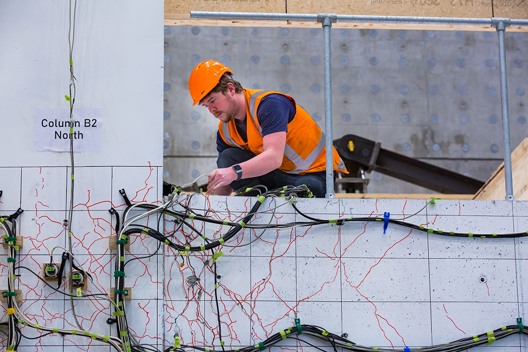 Person wearing high-vis feeding sensor wires into cracked concrete pillar structure.