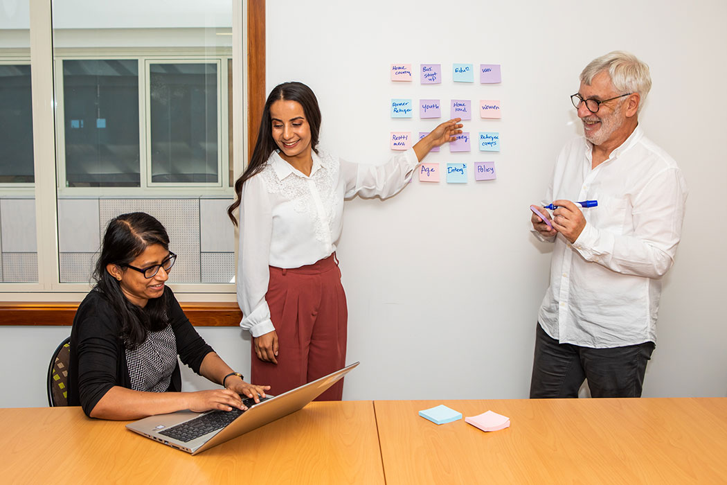 Three people working together, one sitting at a table with a laptop, another putting post notes on a wall, and the other holding a pen and notepad.