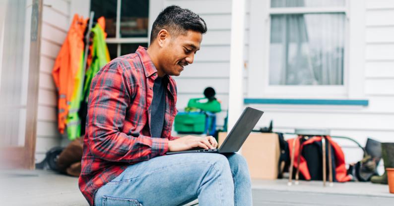 A smiling UC Online student sits on his front porch, resting his laptop on his knees while he completes some online study.