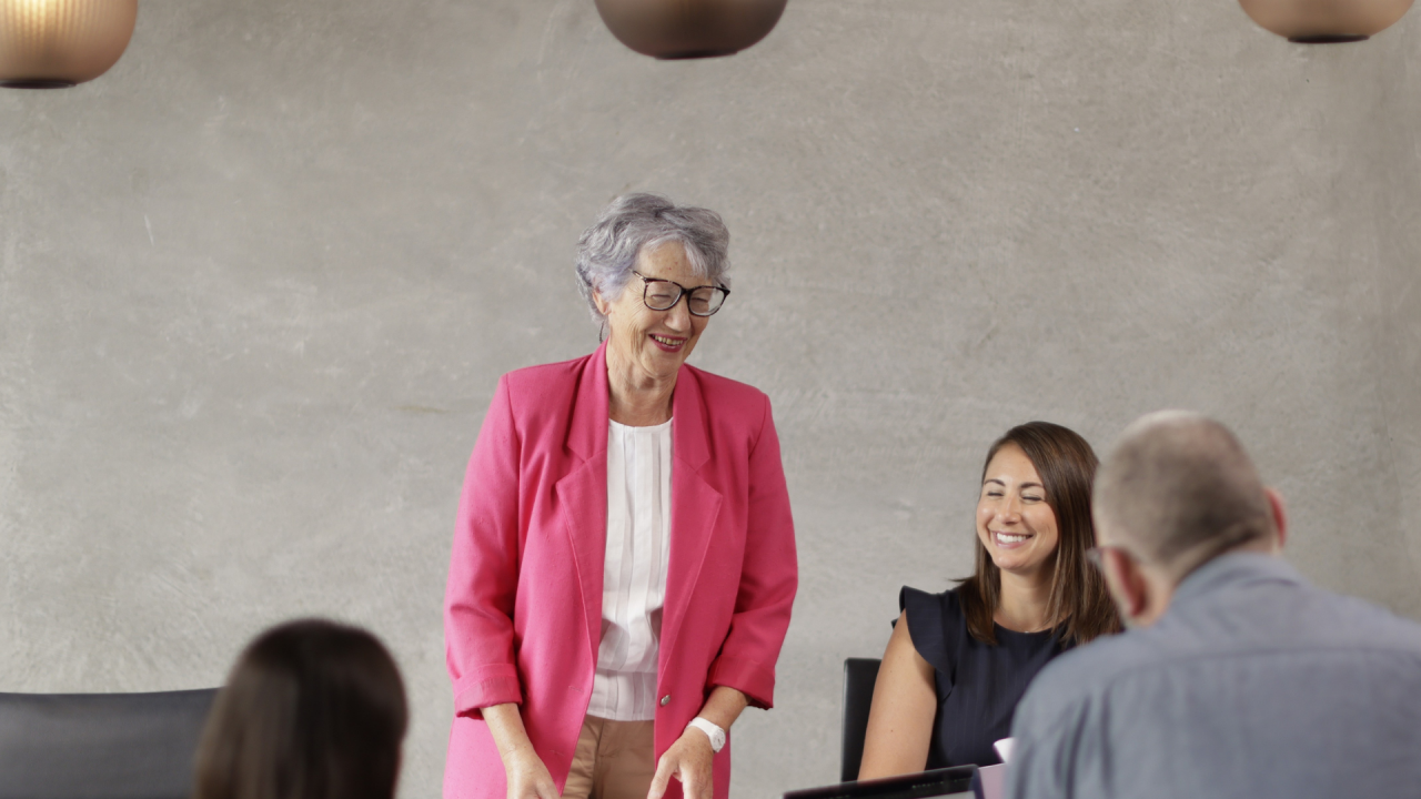 A smiling older woman in a bright pink blazer communicates effectively with colleagues during a productive team meeting.