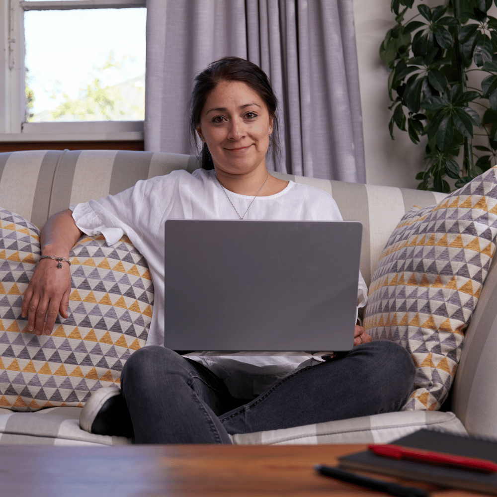 A UC Online student sits cross-legged on a couch with a laptop on her lap. She is relaxed and confident, and smiles at the camera.