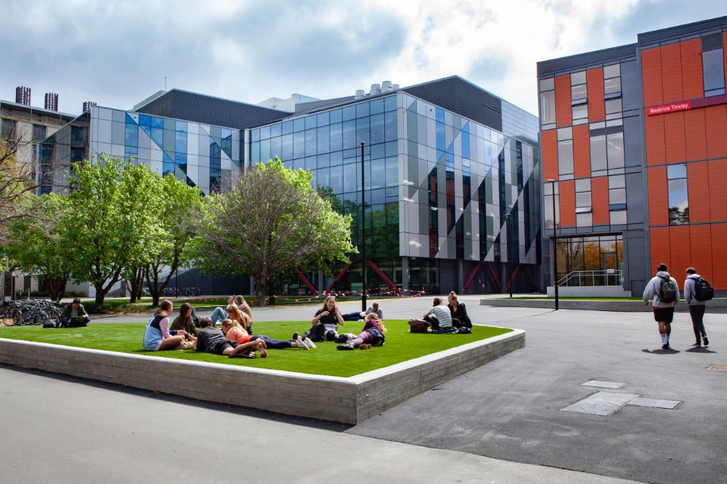 Students sitting on a raised lawn bed in a courtyard, with Rehua and Beatrice Tinsley buildings in the background.