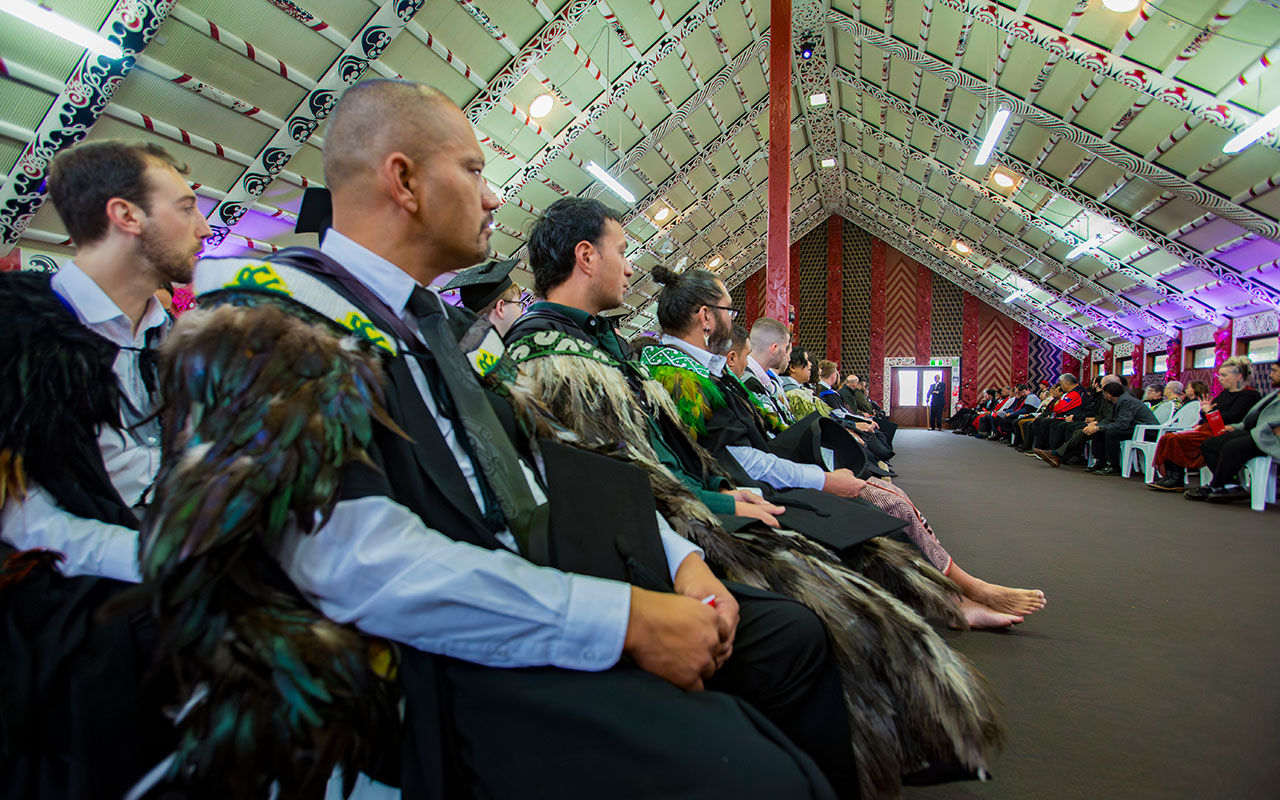 Students wear graduation attire, including Māori korowai, inside a marae.