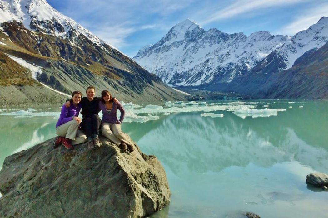 Group of students sitting on rock together with a glacial lake and snowy mountain range behind them.