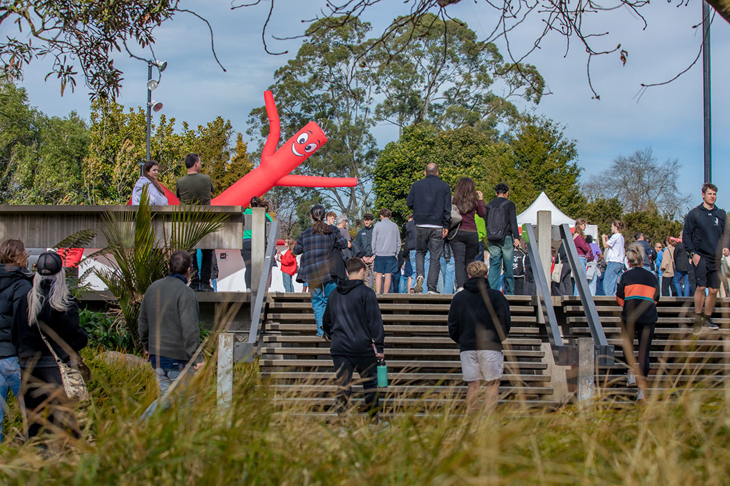A crowd of people walking up the outside steps to the Matariki quad during UC Open Day. A smiling red inflatable-flailing-arm-tube-figure waves above the people.