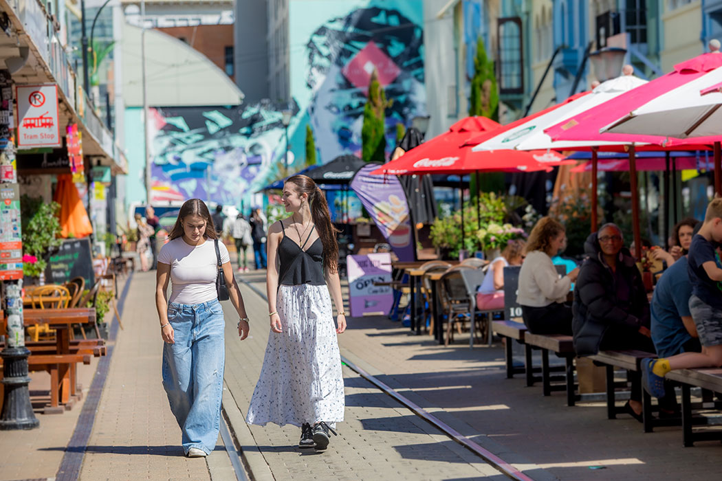 Two UC students walk down the middle of Christchurch's New Regent Street. People can be seen to the side sitting and eating from nearby cafes.
