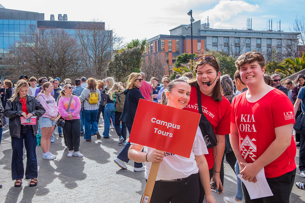 Three UC staff members wearing red t-shirts and holding a sign that reads "campus tours" stand before a crowd of people on UC's campus.