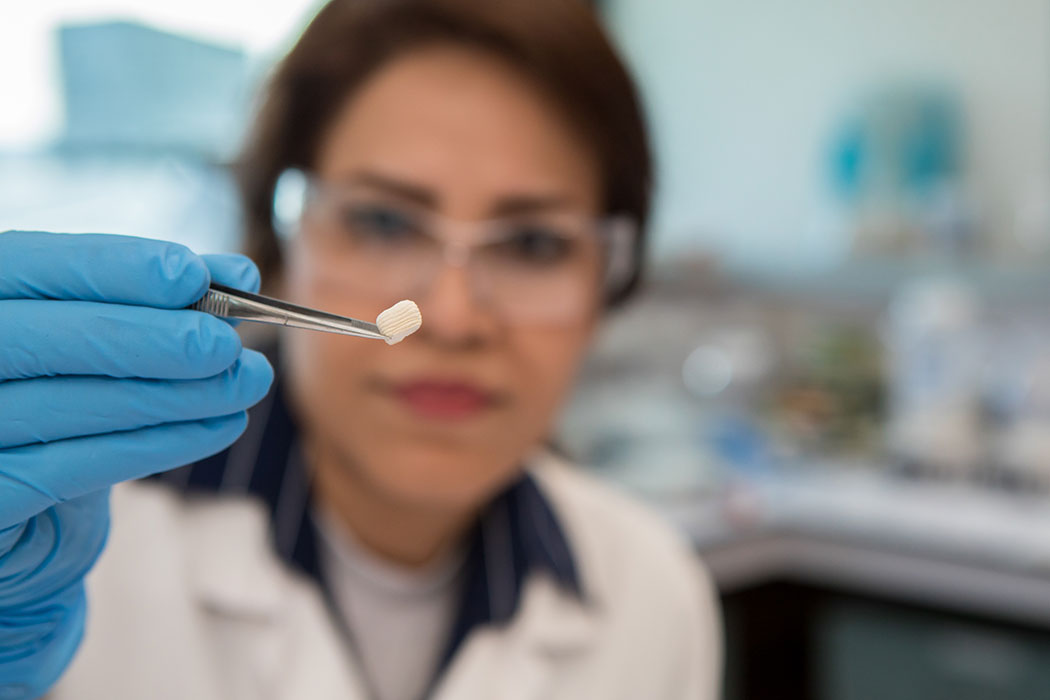 UC researcher Mehrnoush Tangestani wearing lab glasses, coat, and rubber gloves holding a small plastic object with tweezers.