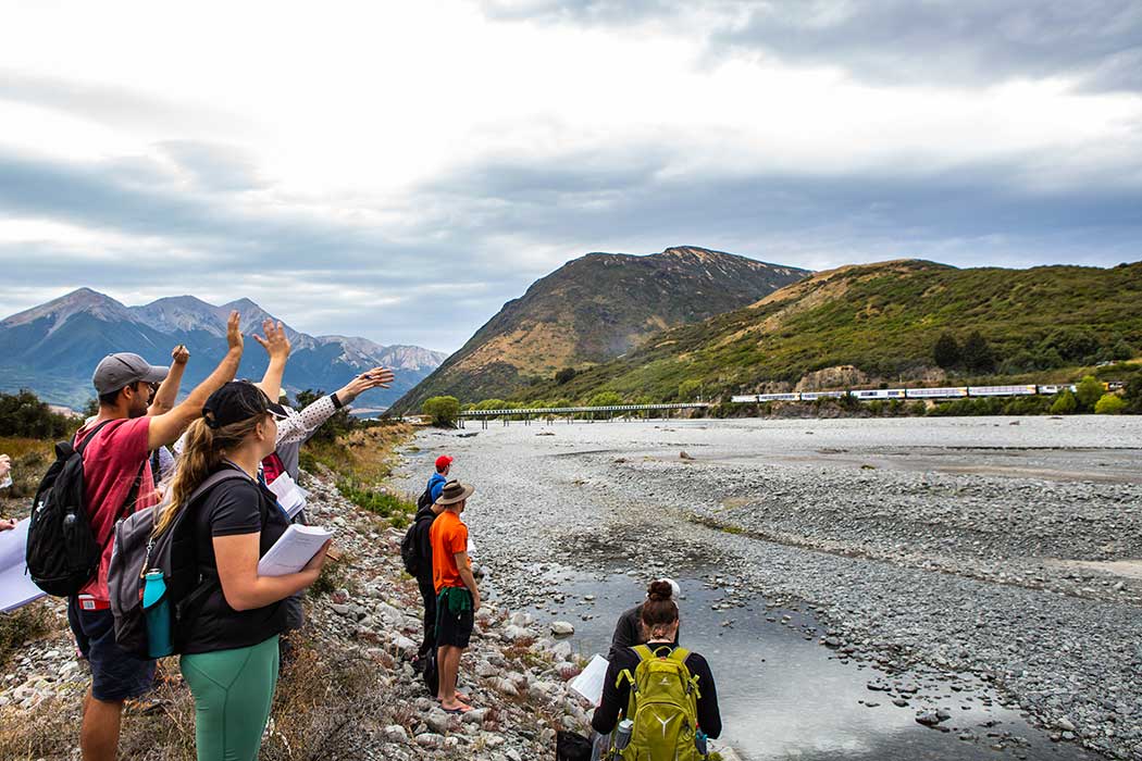 UC students stand on a rocky bank of a river, waving at a passing train on the other side.