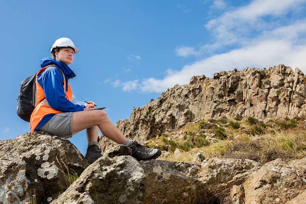 UC student Alex wearing high-vis sits on a large rock and writes on a clipboard. Rapaki Rock formation appears behind them in the background.
