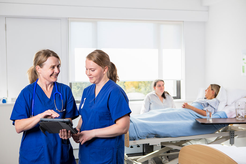 Two UC Nursing students wearing scrubs in a hospital ward setting looking over a patient's file.