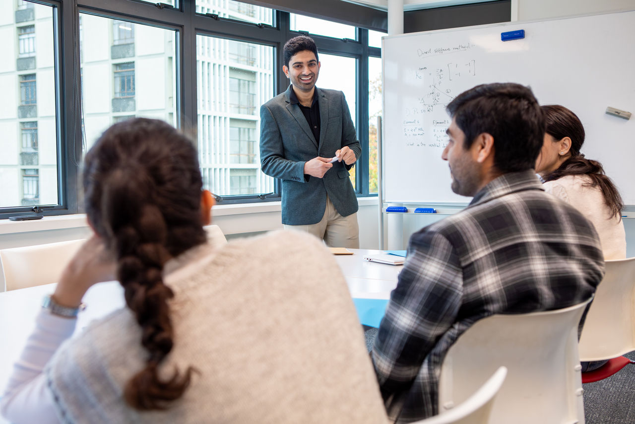 A UC Civil Engineering student presents to a class of students sitting at a table. A whiteboard behind them is covered in formulas.