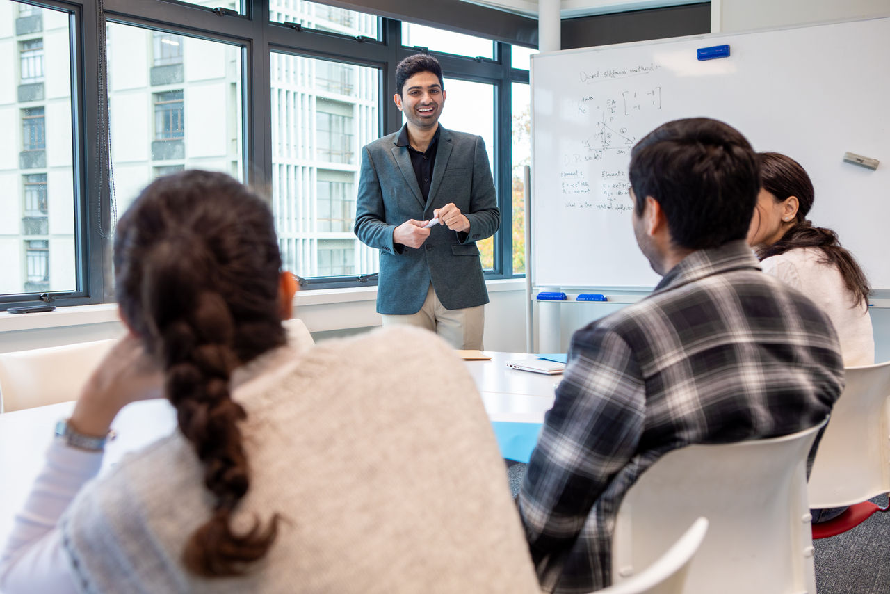 A UC engineer stands at a whiteboard in front of a table of sitting students listening and taking notes.