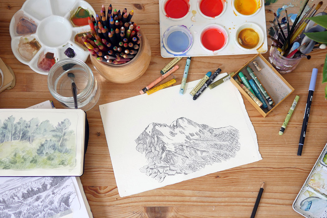 An overhead view of a desk covered in crayons, coloured pencils, paint pots and pallets, and drawings of mountain landscapes.