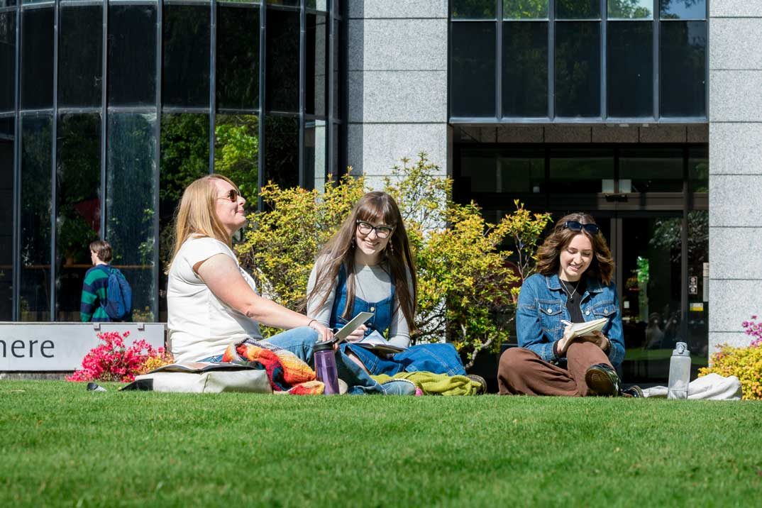 Students sitting together on a lawn outside in the sun, talking and writing notes on notepads.