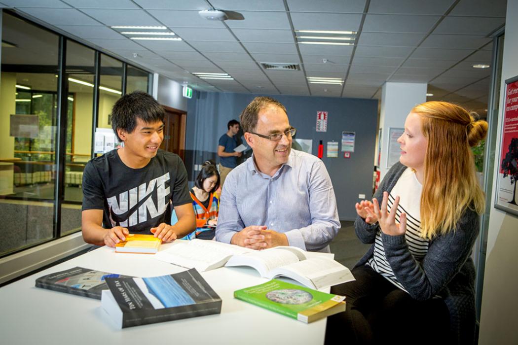 Two students sitting in discussion with a professor with various books on the table in front of them.